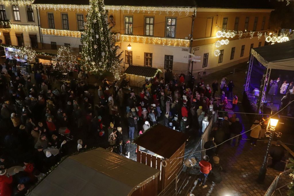 An aerial shot of a crowd in front of a stage, as well as a tall green Christmas tree covered with snowflake ornaments, at Óbuda.