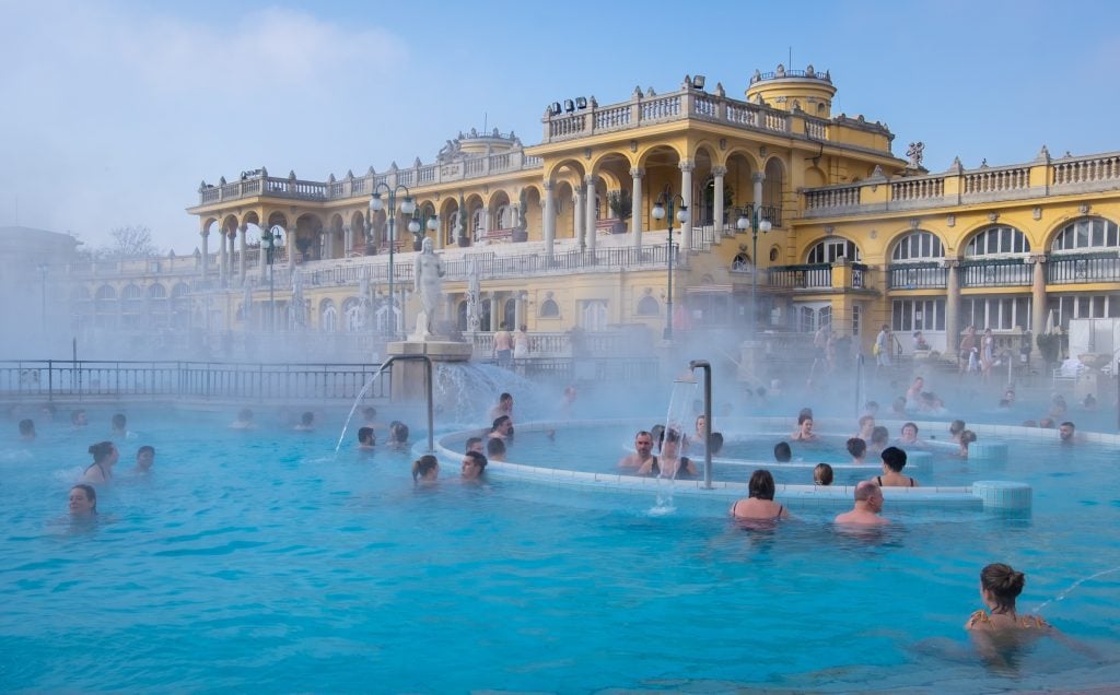 The Szechenyi Baths: an ornate baroque yellow and white building, with people frolicking in the steam-emitting baths in front of it.