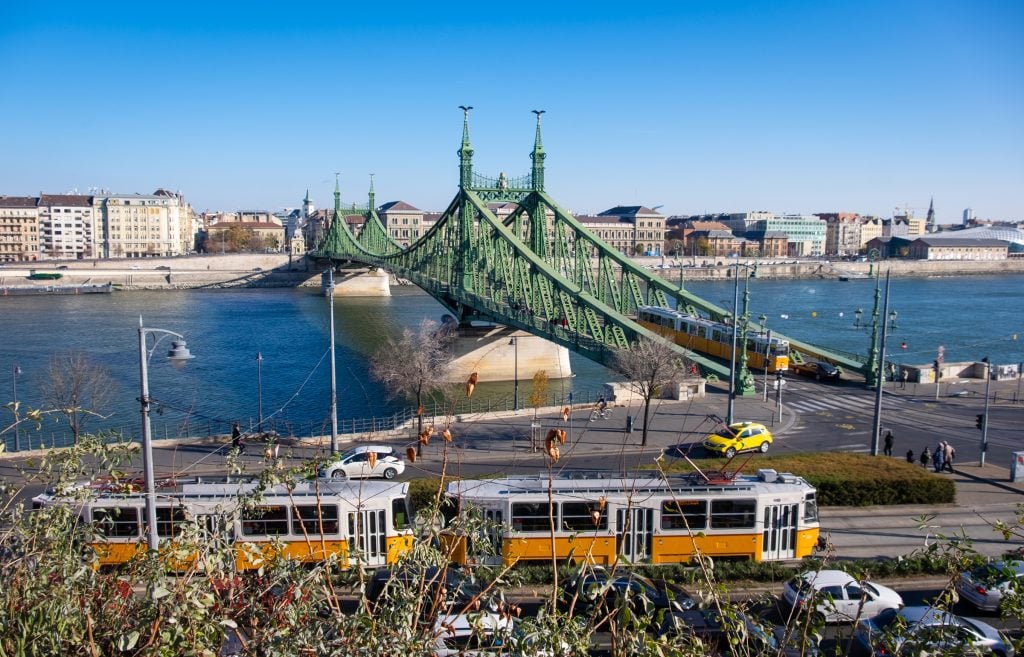 A view of the pointy green bridge of Budapest, spanning the bright blue Danube river, a bright yellow tram on the street in front of it.