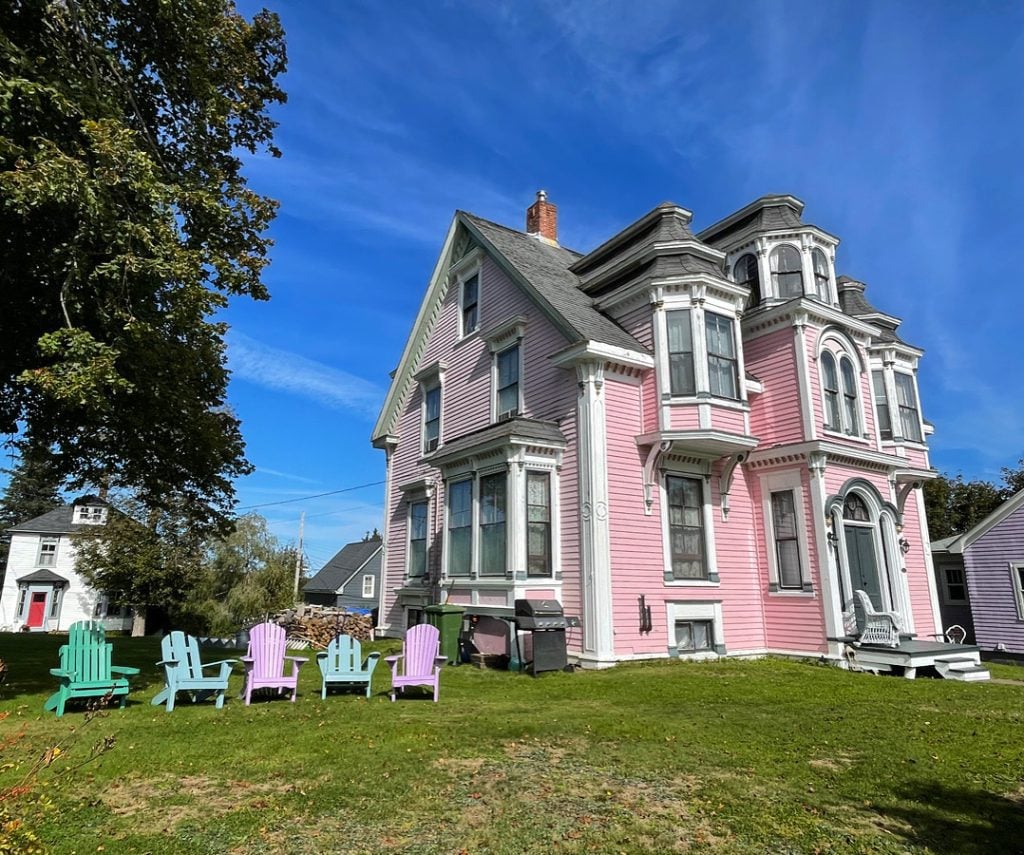 A pale pink Victorian house with lots of turrets and differently shaped pieces popping out, next to a row of pastel-colored Adirondack chairs on the grass.