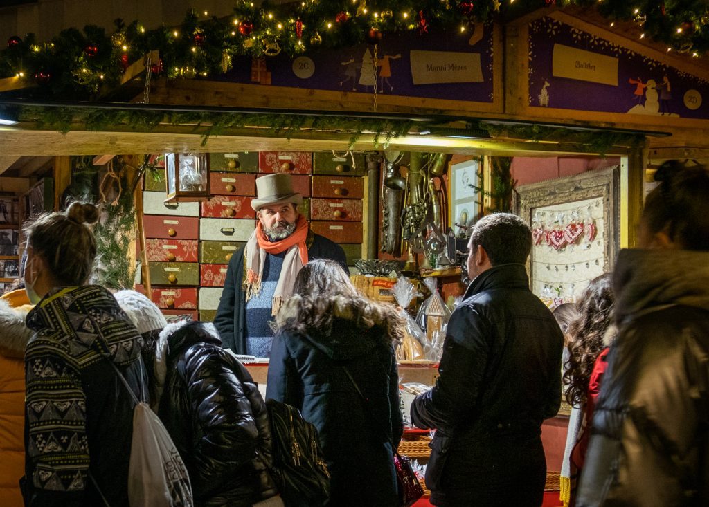 A craft stall at the market, a bearded man selling things while wearing an old-fashioned top hat.