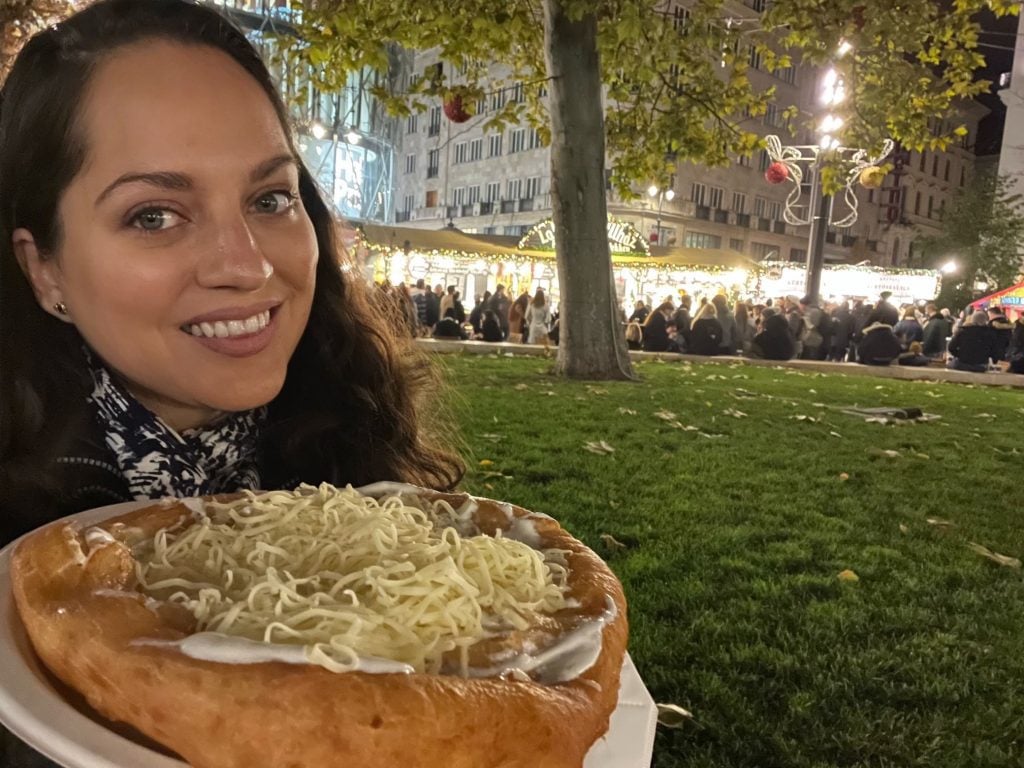 Kate smiling holding a langos: a fried flatbread covered with sour cream and cheese.