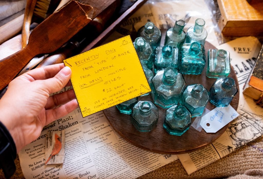 Kate's hand holds a post-it reading "Recently Dug From Tips Upriver from London Inwells, $22 each". Next to her is a wooden platter topped with blue-green octagonal glass inkwells.