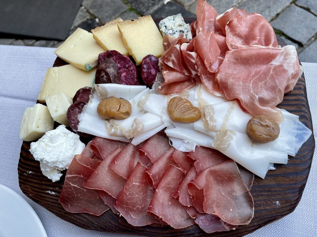 A platter covered with several kinds of sliced meats and cheeses. In the center are slices of white lardo, each topped with a honeyed chestnut.