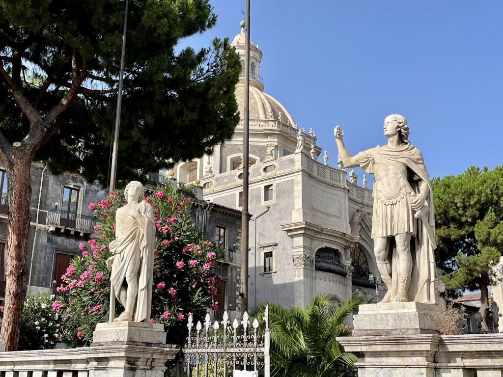 Two statues, a warrior and a woman who looks like she is worshiping him, in front of a fancy dome-topped building in Catania.