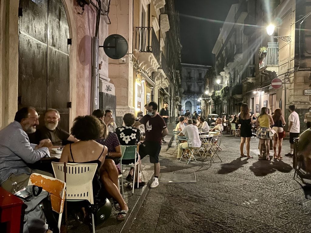 A lively street scene, with a few dozen people sitting at outdoor tables, drinking and having fun, underneath a night sky.