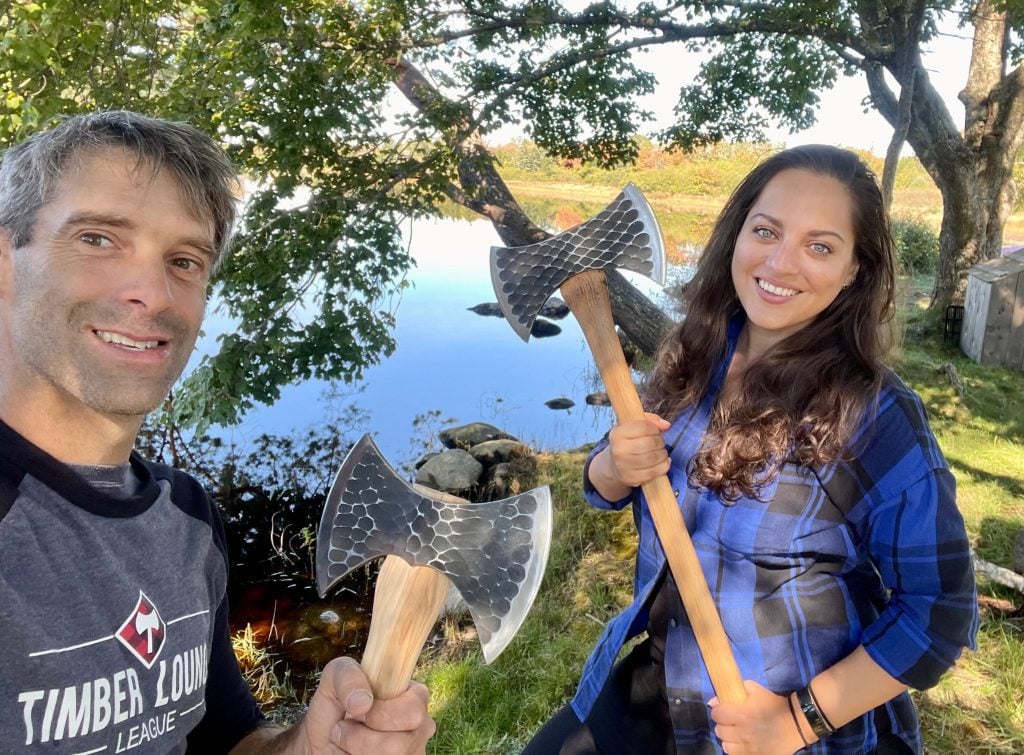 Kate and Darren the Lumberjack hold giant axes and grin at the camera. Kate wears a black and blue plaid flannel shirt.