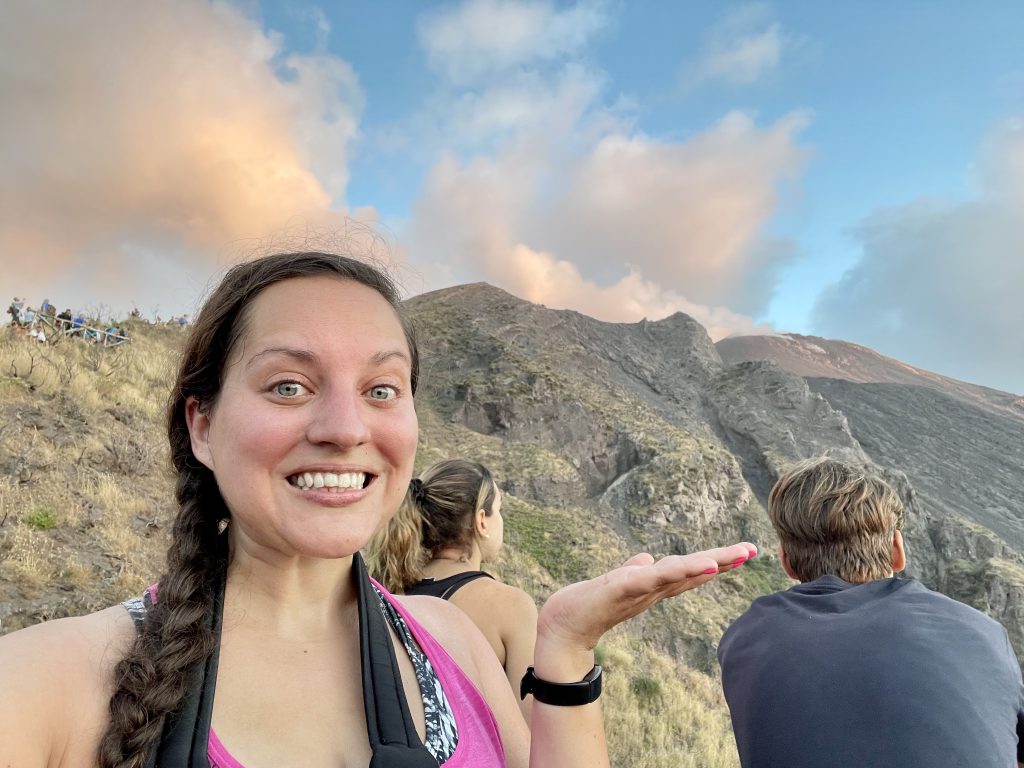 Kate standing in front of the volcano of Stromboli, holding her hand out and grimacing. Her hair is pulled into a side braid.