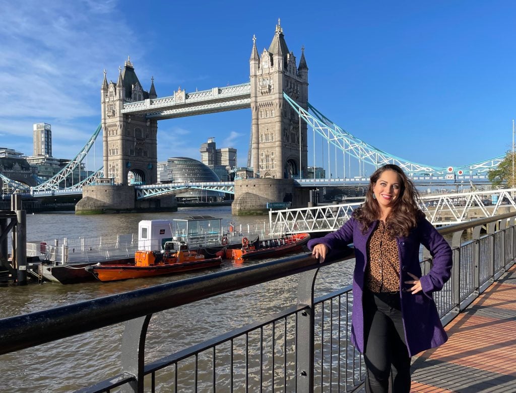 Kate standing in front of the Tower Bridge in London, wearing a purple coat and a leopard print blouse.