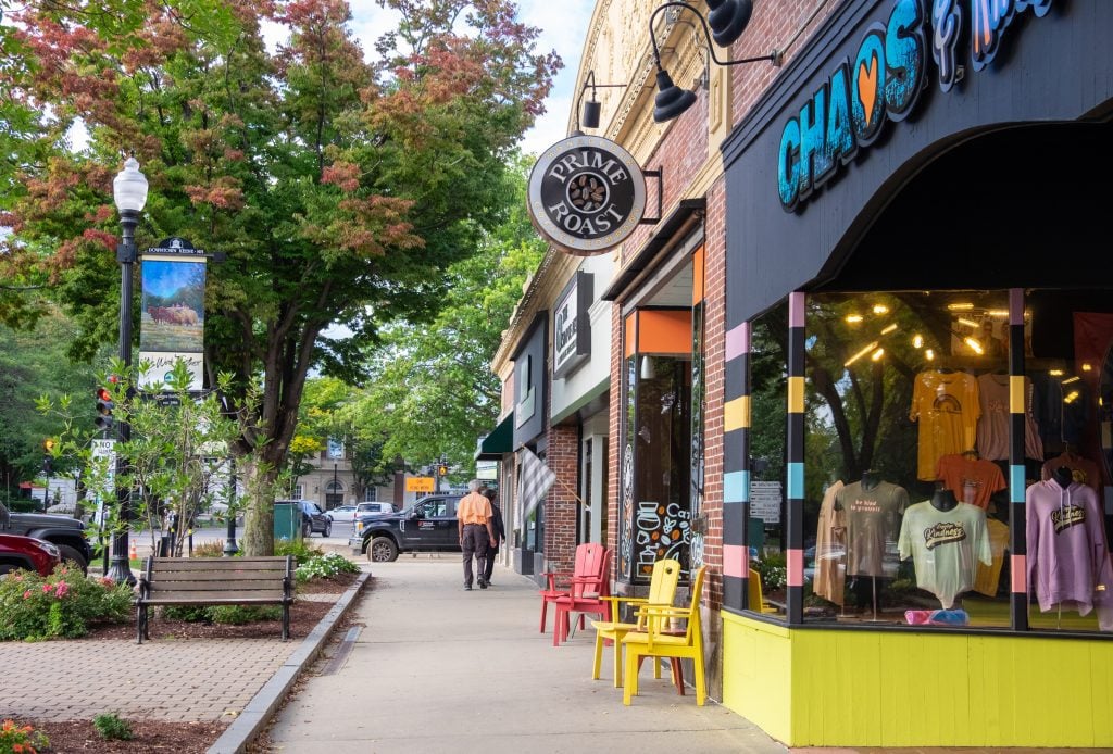 A sidewalk in downtown Keene, New Hampshire, next to a t-shirt shop and a coffeeshop. One tree is starting to turn red and orange.