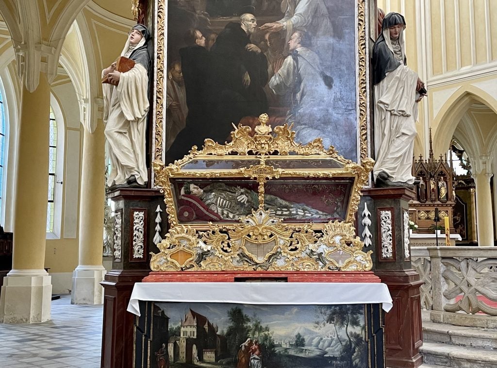 Inside a church, an ornate golden glass coffin, with a sculpture of a skeleton inside.