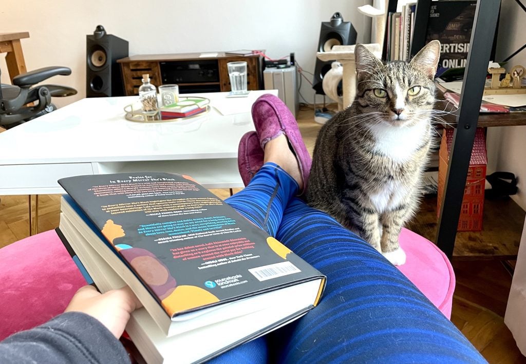 Kate holding a book on her lap with her feet up in blue leggings. Lewis the cat sits right next to her legs on the pink ottoman and glares at the camera.