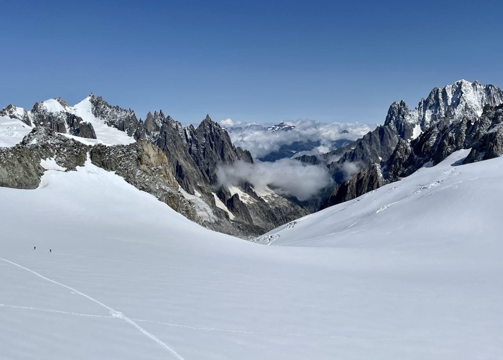 Snow in the Alps in August, jagged gray mountains in the distance. There are two tiny people that look like ants, which shows you how big the landscape is.