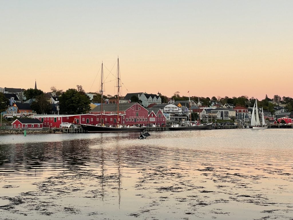 A sunset over Lunenberg's harbor. You see a bright red warehouse-type building, a tall ship, and rows and rows of seaside homes. The sky is pink and faded.