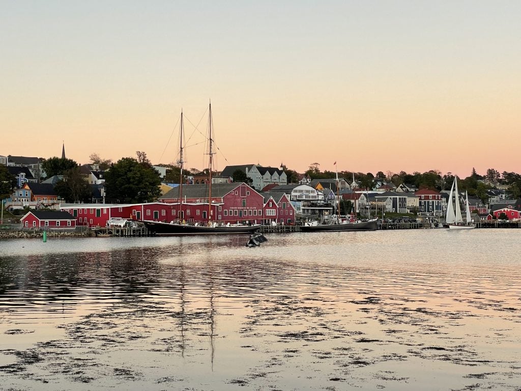 A sunset over Lunenberg's harbor. You see a bright red warehouse-type building, a tall ship, and rows and rows of seaside homes. The sky is pink and faded.