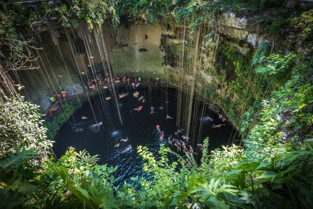 A very deep circular cenote with dozens of people swimming inside it, vines hanging in it.