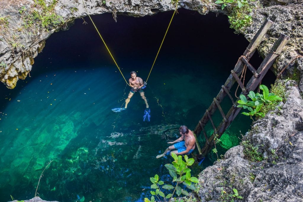 Two men snorkeling in flippers, one sitting on the bottom of a ladder leading into bright blue-green cenote.