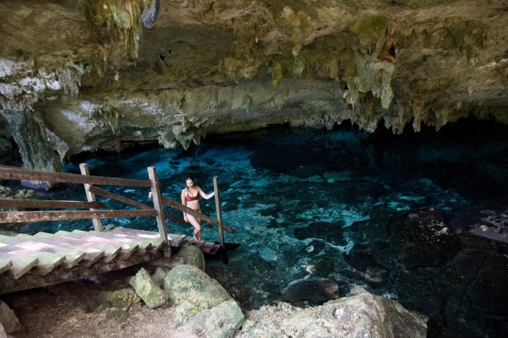A girl in a bikini standing at the base of a wooden flight of stairs leading into a dark teal cenote surrounded by rocks.