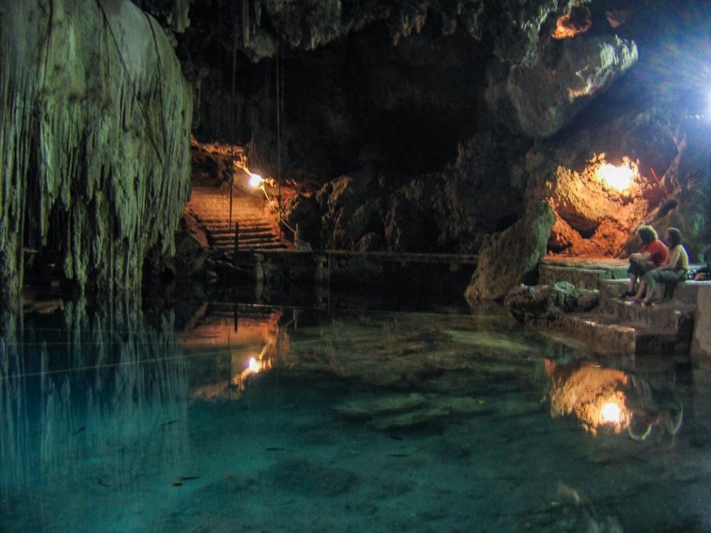 Two women sitting on the edge of a dark, clear cenote inside a cave.