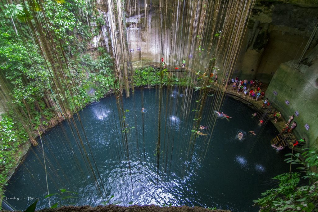 A large round cenote with people swimming in the navy blue water and several skinny vines hanging inside.