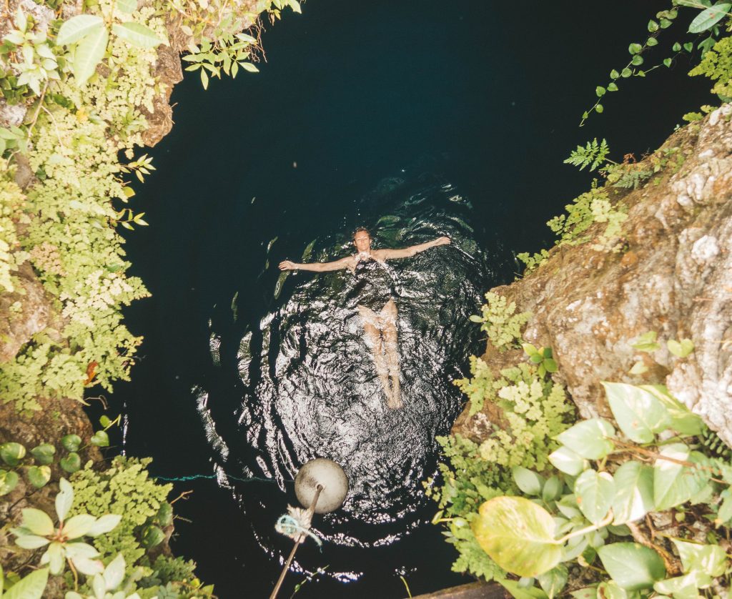 A woman in a black bathing suit swimming on her back in a dark green cenote, surrounded by cave walls and plants.