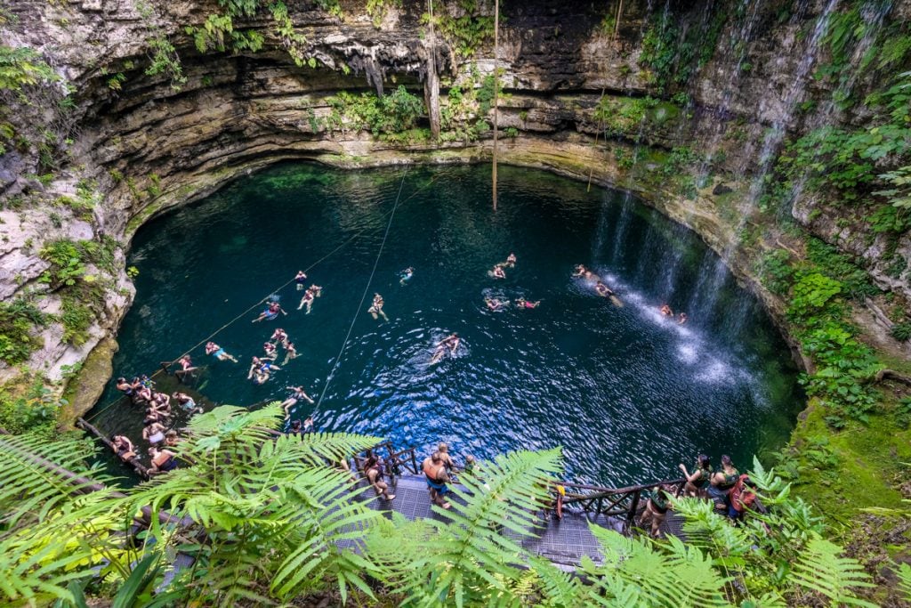 A round cenote with about a dozen people swimming inside it, and some light waterfalls falling on one side, all surrounded by lush vegetation.