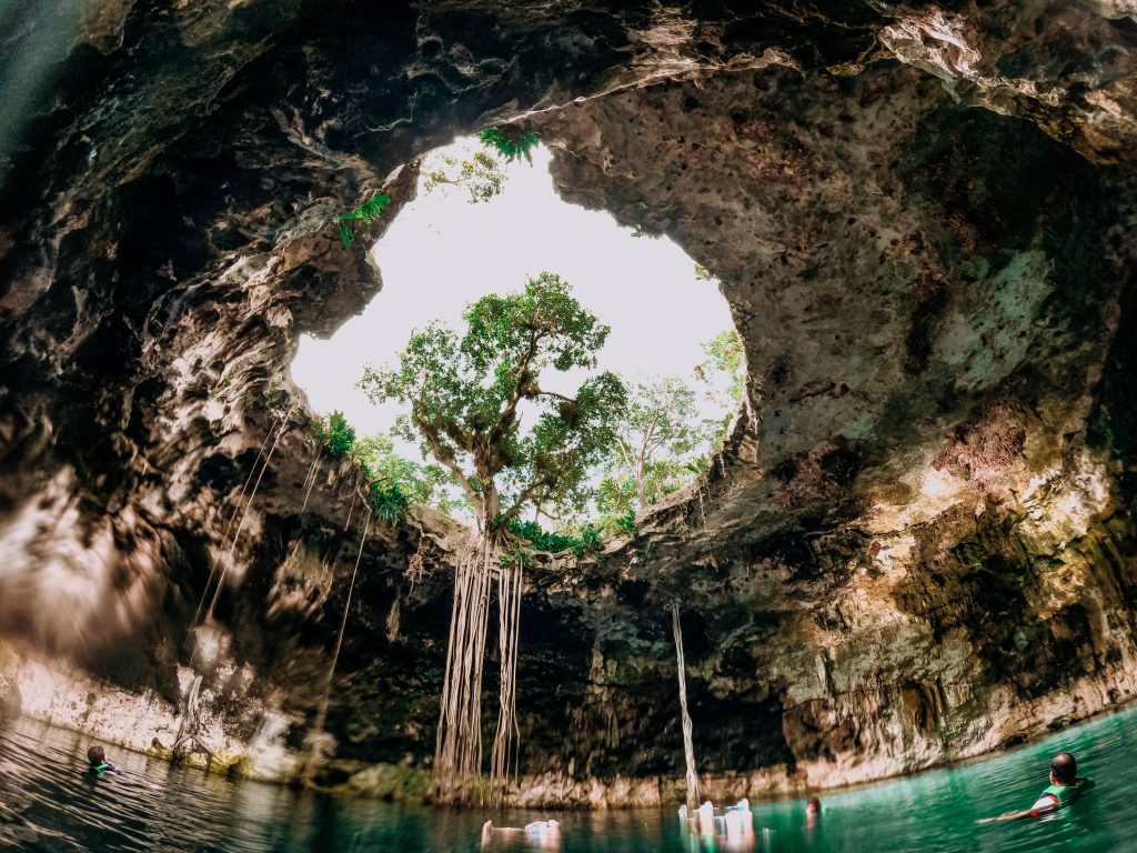 A bright green cenote that looks like a cave, with a circular opening at the top that lets light in.