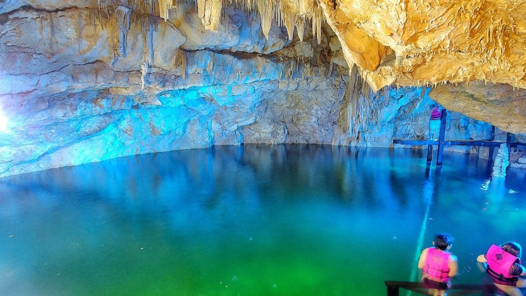 Two kids with bright pink life jackets in front of a placid, clear blue-green cenote inside a cave.