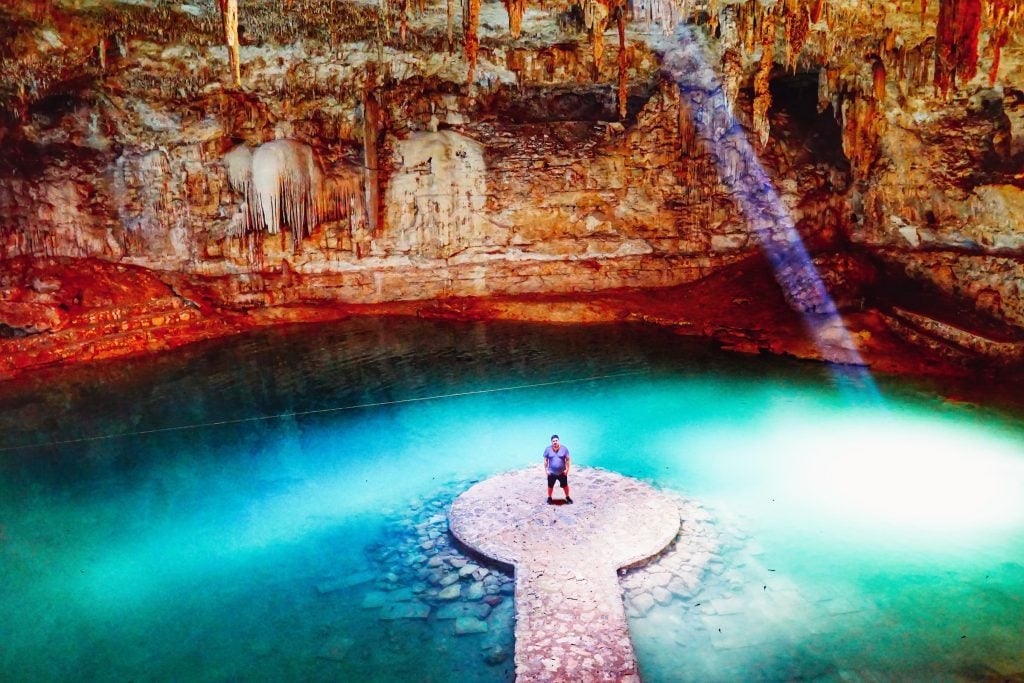 Nathan standing on a round platform in the middle of bright turquoise water, surrounded by cave walls, rays of sunlight coming into the cenote from overhead.