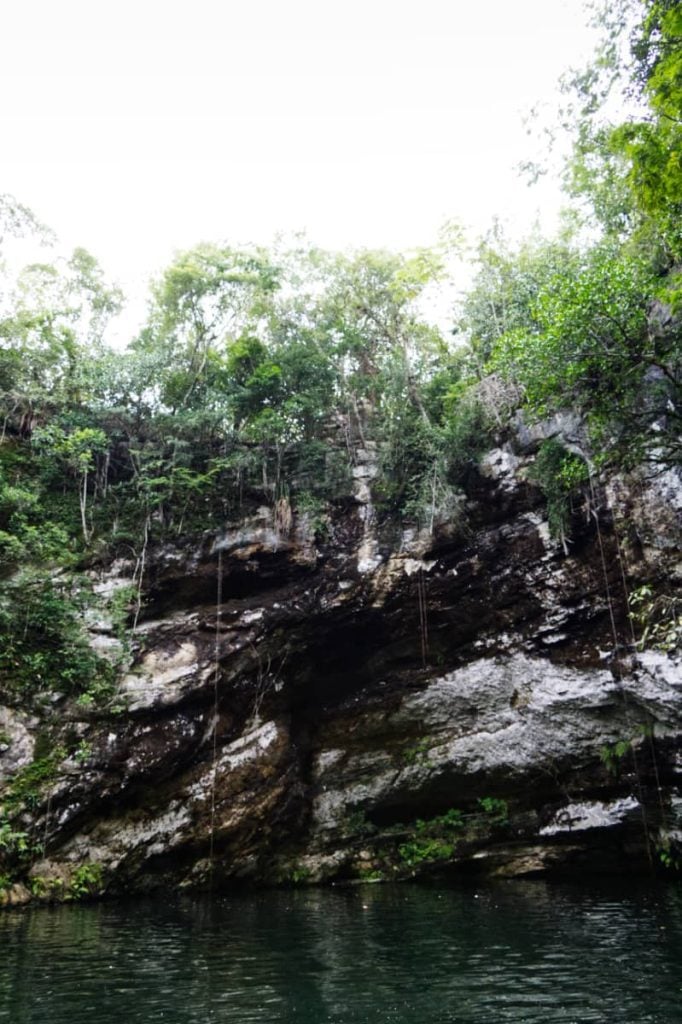 Tall gray cave walls in front of a pool of dark green water, almost black.