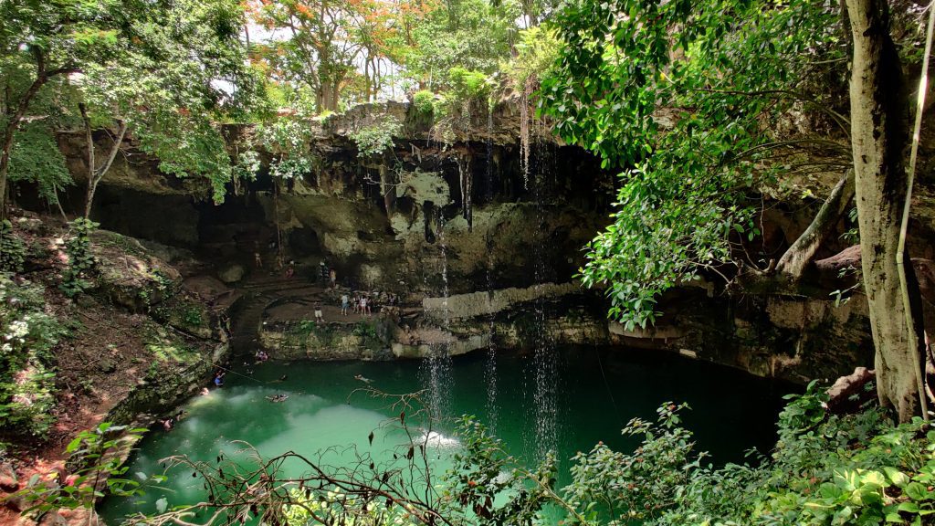 An enormous outdoor cenote with jade green water, light waterfalls falling into it from above, surrounded by trees and plants.