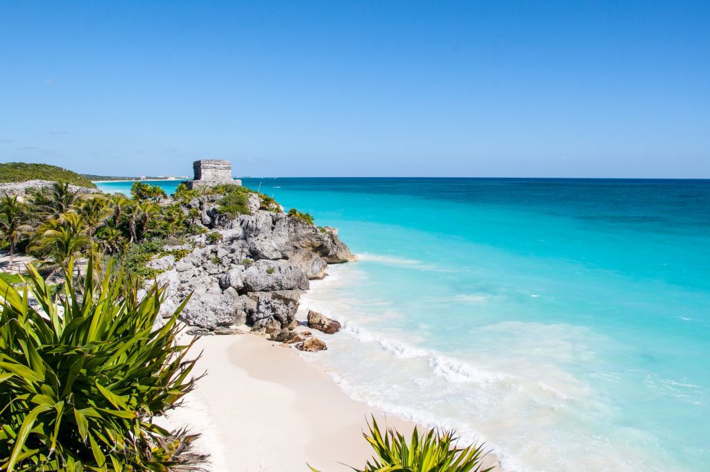 The Tulum Mayan ruins, set on a rocky cliff next to a white sand beach and bright blue ocean.
