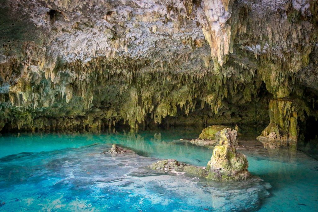 A cave with lots of stalactites filled with bright aqua water.