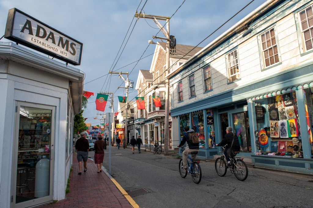 Shop-lined Commercial STreet, Portuguese flags hanging across the street, two people biking down it.