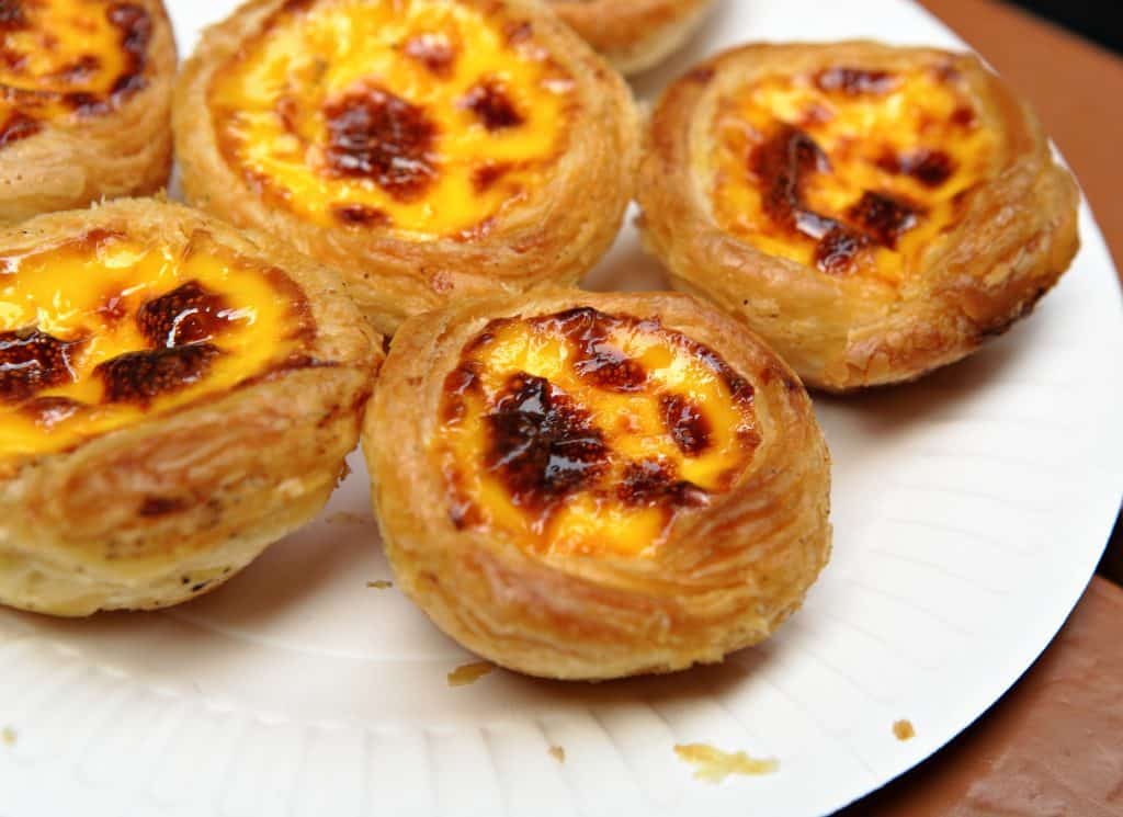 Several small flaky yellow Portuguese egg tarts on a paper plate. You can feel the grease just looking at them.