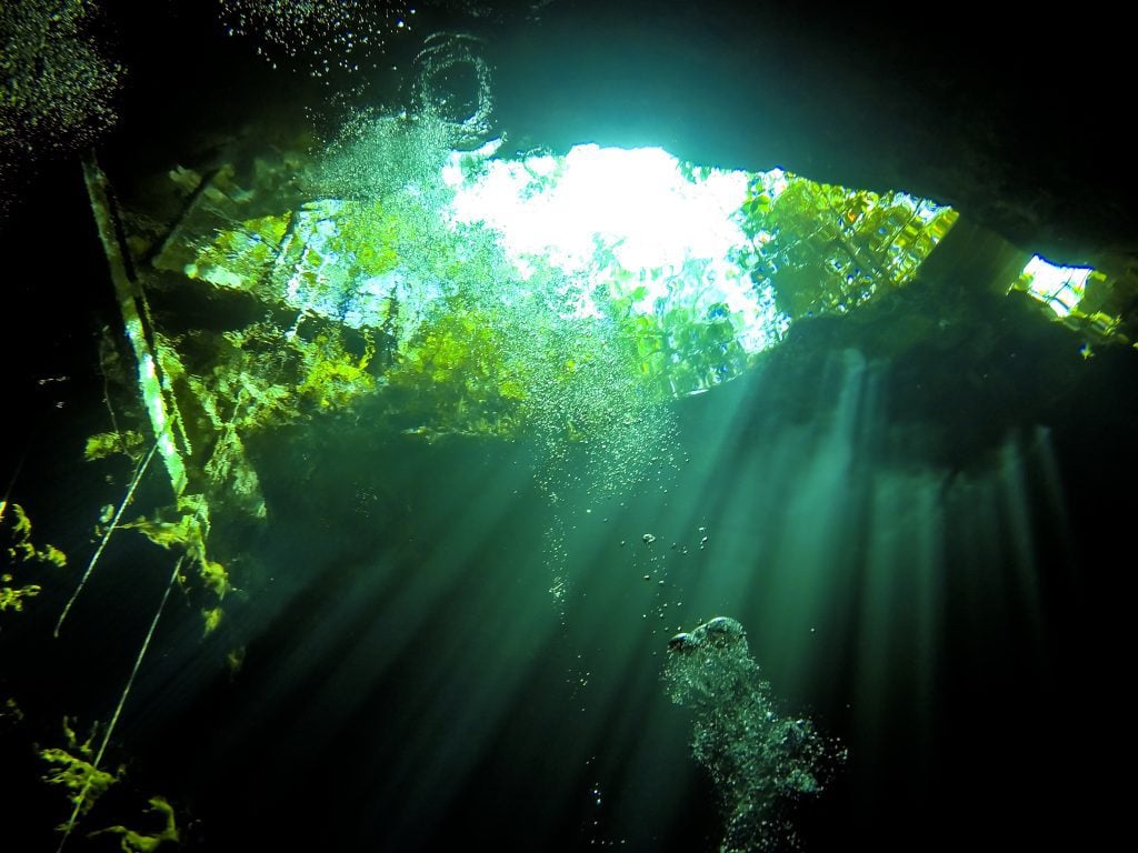 An underwater shot of a cenote, inside a cave, rays of sunshine and water bubbles floating through.