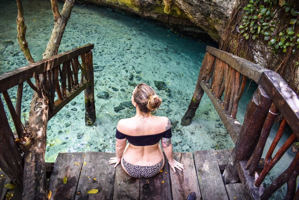 A blonde girl in a black bikini top and leopard bottom sitting on a platform leading into a sea foam green cenote.