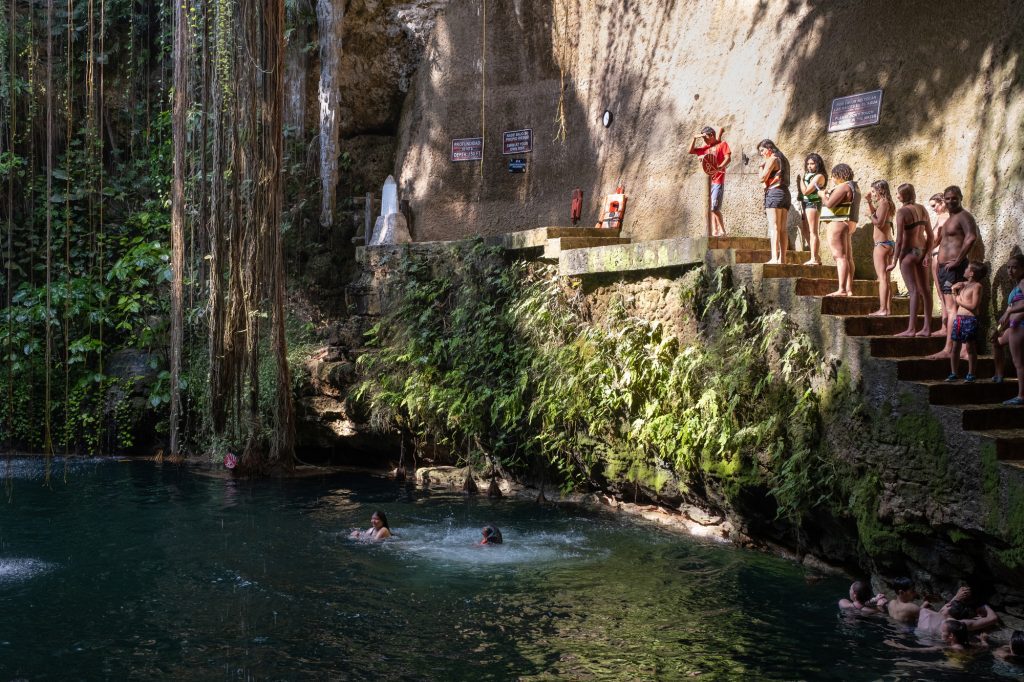 A group of people standing on a staircase, in line to jump on the rope swing that goes into a cenote.