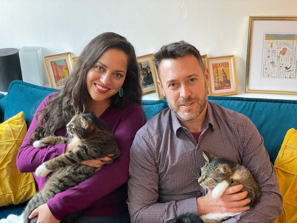 Kate and Charlie sitting on a teal couch, each holding a gray cat in their arms, smiling.