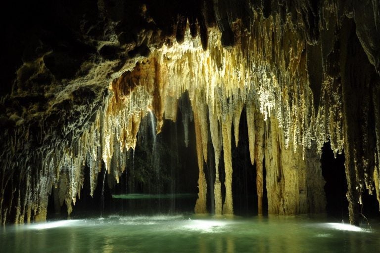 Long, skinny stalactites hanging into a pale green pool inside a cenote.