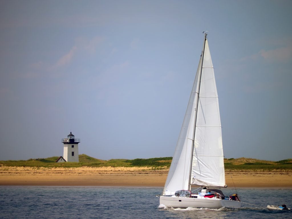 A sailboat in the water at Provincetown, the white Wood End Lighthouse on shore in the background.
