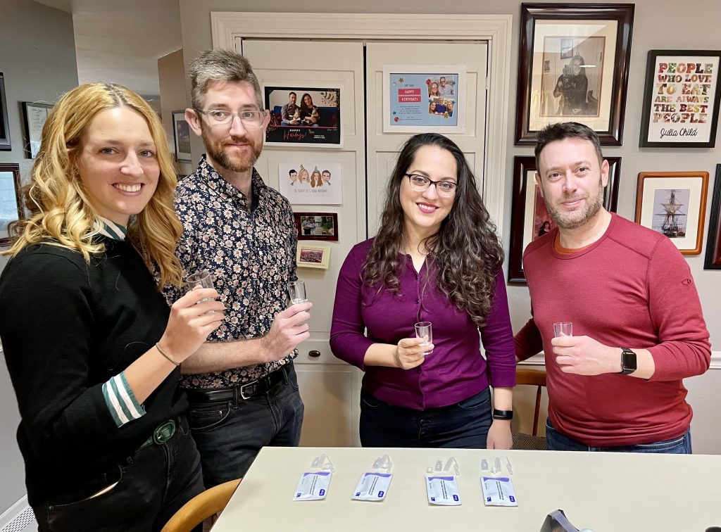 Kate, Charlie, Kate's sister Sarah and her husband Matt, standing in a kitchen, holding shot glasses, and standing in front of rapid tests.
