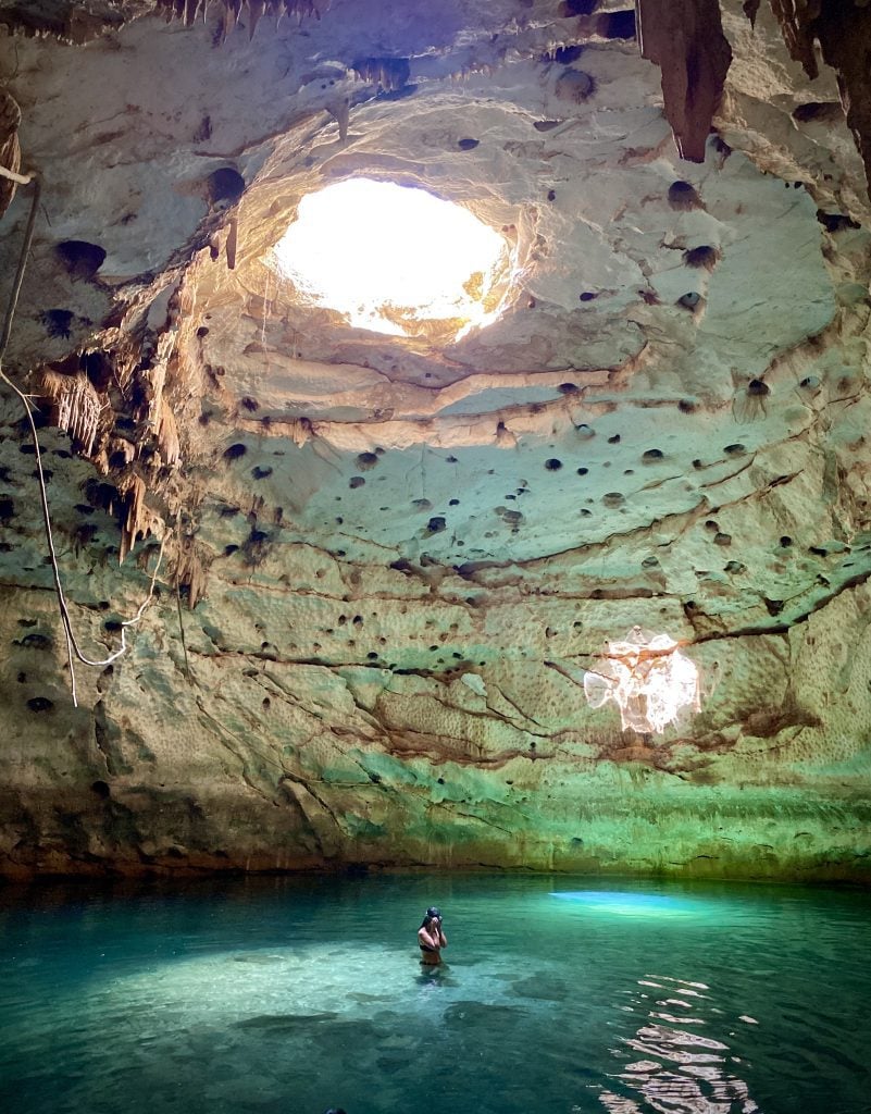 A woman standing in a cenote cave, a large opening in the rock illuminating her from above.