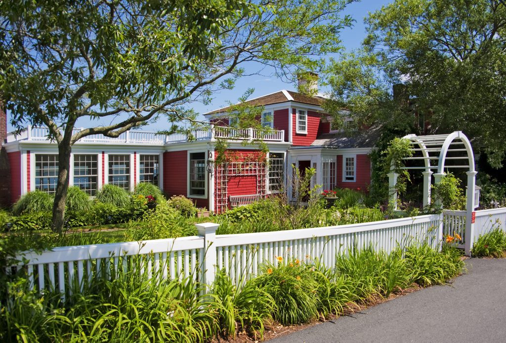 A large red wooden house with white trim, a white picket fence in front, lots of overgrown wildflowers around it.