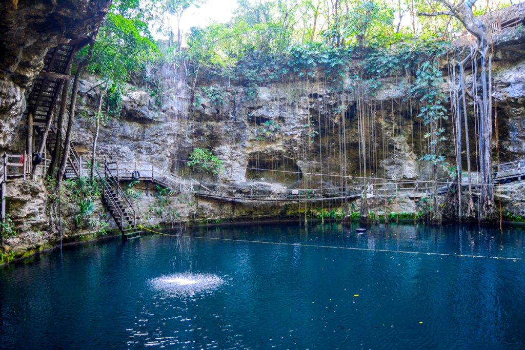 A dark blue cenote surrounded by cave walls, a wooden platform circling the edge, several skinny vines hanging into it.