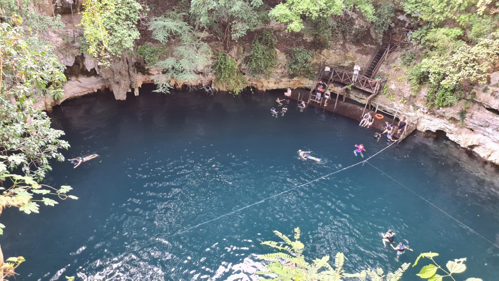 An open cenote looking like a teal lake, a few people swimming inside it next to a wooden platform.