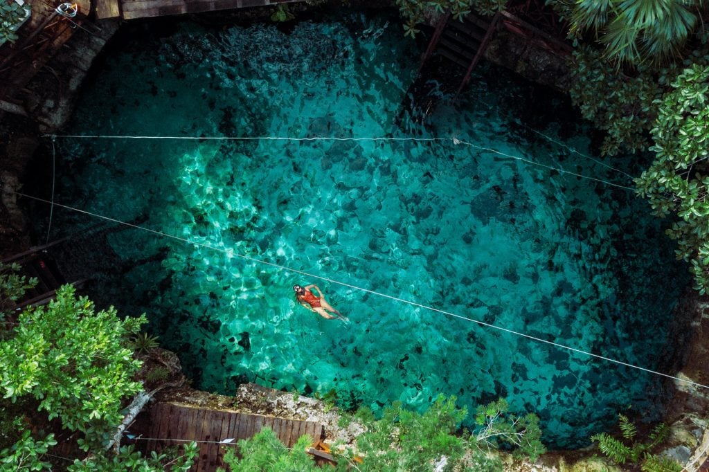 An aerial drone shot of a girl in a red bathing suit swimming in a clear, bright teal cenote surrounded by lush vegetation.