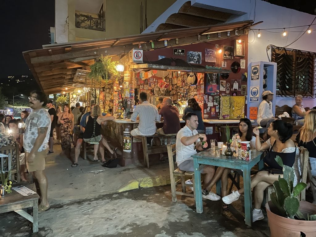 An outdoor bar in Sayulita, Mexico, covered with hundreds of stickers. Some people sit at outdoor tables, some people sit on swings hanging on the edge of the bar.