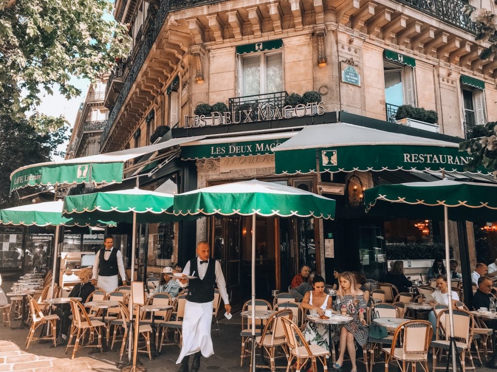 Cafe Les Deux Magots on a corner in Paris, with green umbrellas, tuxedoed waiters, and people dining at small tables outside.