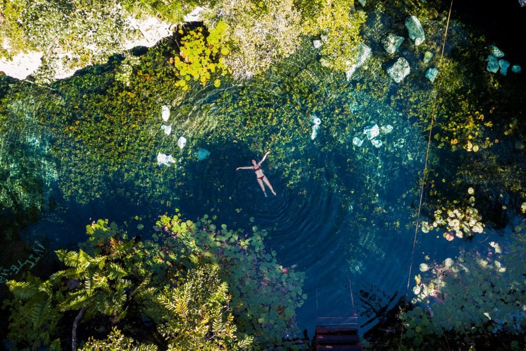 An aerial drone shot of a girl in a bikini swimming in a dark green cenote, surrounded by rocks and plants.